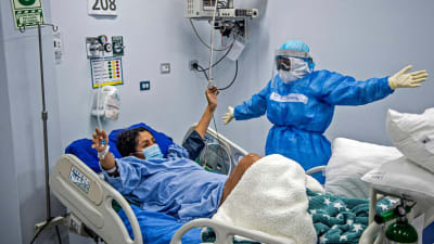 En sjukskötare i skyddsutrustning står vid en patients säng. De håller båda ut händerna åt sidorna.