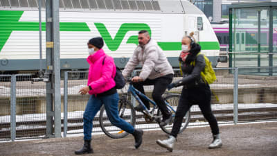 Två kvinnor i munskydd promenerar och en man utan munskydd på cykel på Helsingfors järnvägsstation 25.3.2021