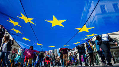 Människor går på gata och håller i en enorm EU-flagga. Bilden från under flaggan.