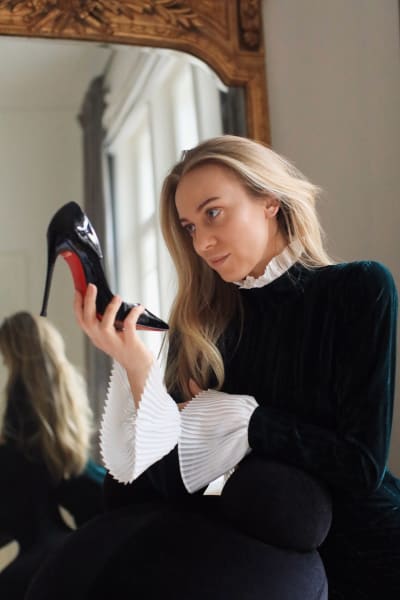 På bilden syns influeraren Sandra Hagelstam som håller i och tittar på en svart högklackad sko med röd sula.