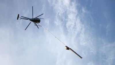 Helikopter flyger i luften. Under helikoptern hänger ett sågblad.