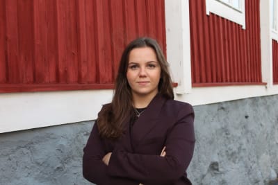 En kvinna som står med armarna i kors utanför ett trähus. 
