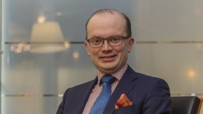Penna Urrilla i en blå kostym med en röd och vitrandig skjorta. Han sitter i en skinnfotölj.