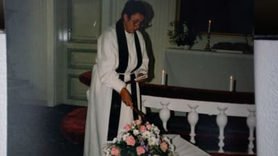 En kvinnlig präst tänder stearinljus som står på ett vitt bord med rödfärgade blommor i en vas.