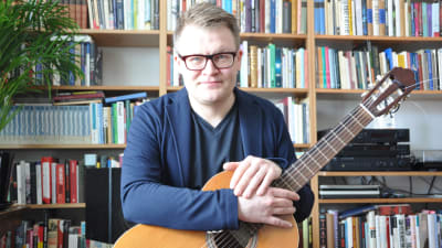Forskaren Juha Torvinen sitter i sitt vardagsrum med gitarren i knät