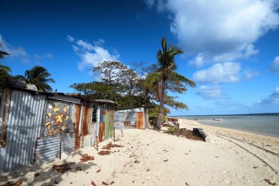 På bilden syns strandhytter, palmer och blå himmel på ön Masig som ligger i Torressundet norr om Australien.