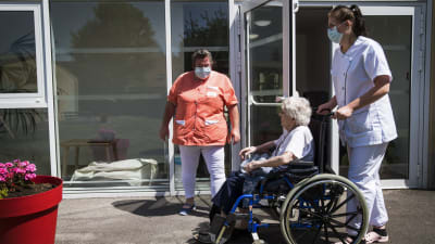 Två sjukskötare tar hand om en äldre kvinna i en rullstol utanför ett äldreboende.