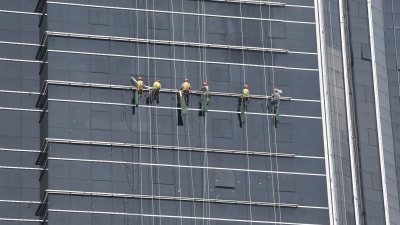 Sex arbetare med hjälm står på en ställning på en hög glasklädd fasad.