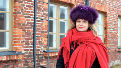 Kvinna i röd sjal och lila pälsmössa utanför rödtegelhus.