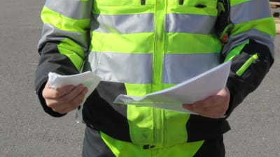 En man i svarta och neongula arbetskläder håller ett munskydd i ena handen och en bunt med papper i andra handen. Man ser inte ansiktet på mannen.