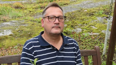 En man i pikeskjorta i mörkblått med vita ränder sitter i en trädgårdsgunga. Han ser allvarlig ut.