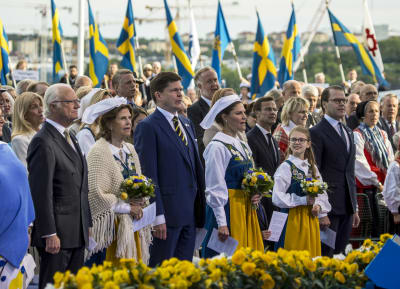 Kung Carl XVI Gustaf, drottning Silvia, riksdagens talman Andreas Norlén, kronprinsessan Victoria, prinsessan Estelle och prins Daniel firar Sveriges nationaldag på Skansen i Stockholm. Bakom dem vajar en rad svenska flaggor.