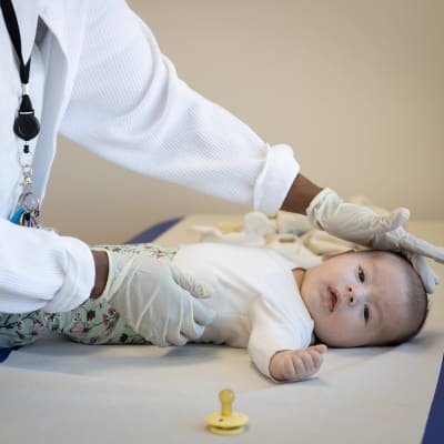 En baby ligger på ett skötbord och tittar in i kameran. En skötare masserar babyns huvud med handen.