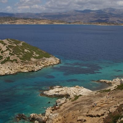 Kartion muotoinen pikkusaari sinisessä meressä, edustajja isomman saaren kalliorantaa.