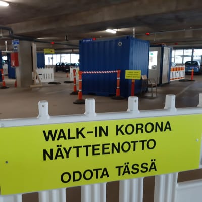 En gul skylt med texten Walk-in korona, fäst på ett staket vid en coronatestning.