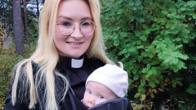 En kvinna med blont, långt hår och glasögon, en svart kappa, prästkrage och en bebis i en bärsele på magen.