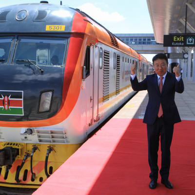 Kiinan suurlähettiläs nostaa peukkuja junan vieressä.