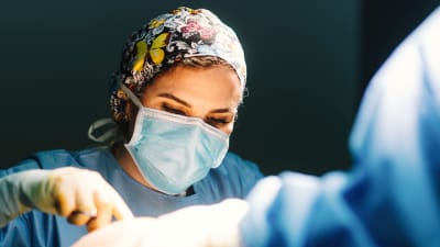 En kirurg i musnkydd håller i ett verktyg och tittar ner.