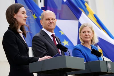 Sanna Marin, Olaf Scholz och magdalena Andersson håller presskonferens bakom var sitt podie i blåsiga förhållanden. I bakgrunden ländernas flaggor.
