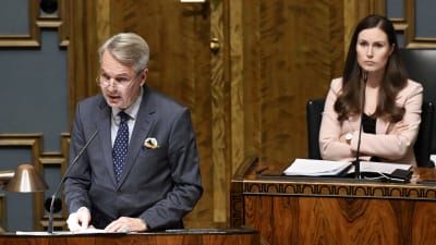 Pekka Haavisto står vid ett podium i riksdagen. I bakgrunden syns statsminister Sanna Marin som har armarna i kors.