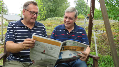Två män sitter i en trädgårdsgunga och tittar i en papperstidning, Hangötidningen. Sommar, utomhus.