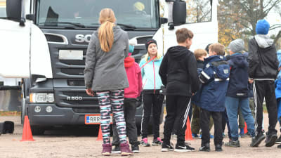 En grupp barn köar utanför en vit lastbil på en grusplan. En flicka med blont  hår och mössa står vänd mot kameran, resten har ryggen till.