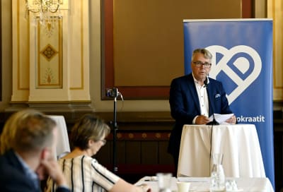 Peter Östman står och talar framför Kristdemokraternas banderoll, Sari Essayah lyssnar i förgrunden.