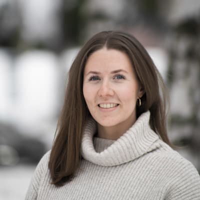 En kvinna ser in i kameran. Hon ser glad ut. I bakgrunden träd.