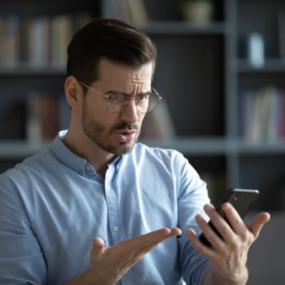 Man med glasögon tittar argt på sin telefon och slår ut med händerna 