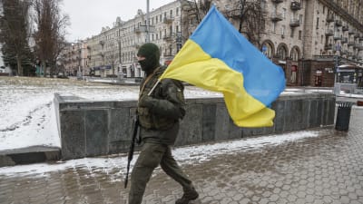 En soldat i militärutrustning går med ett vapen i ena handen och Ukrainas flagga i den andra. I bakgrunden en gammal byggnad.