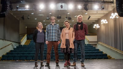 Två kvinnor och två män står på en scen och tittar in i kameran. Bakom dem skymtar en åskådarläktare och flera strålkastare.