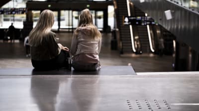 Två flickor med långt hår sitter med ryggen mot kameran på en bänk på köpcentret Tripla i Helsingfors.
