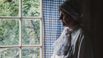 Kvinna i vit spetsklänning och vit klut på huvudet tittar ut genom fönster, som omgärdas av blåvitrutig gardin.