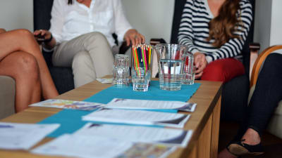 Ett bord med vattenglas, en vattenkanna och en bunt pennor. I bakgrunden syns fyra personer som diskuterar med varandra.
