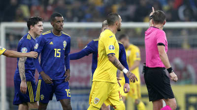 Svenska och rumänska fotbollsspelare följer efter domaren som pekar åt sidan.