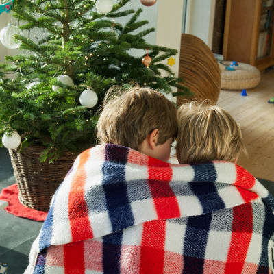 Två små barn sitter under samma filt framför en liten julgran.