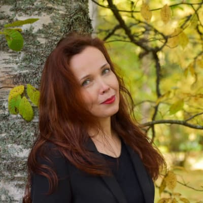 Pitkätukkainen nainen katsoo hymyillen kameraan ja nojaa syksyiseen koivunrunkoon.