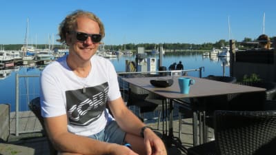 En man med blont lockigt hår, solglasögon, t-skjorta och shorts sitter på en uteservering vid en hamn. (Ekenäs gästhamn)