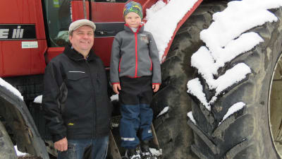 En man med blå jacka och grå keps står framför en stor röd traktor (skogstraktor). På traktorns trappsteg står en sexårig pojke. De ser in i kameran. Snö på traktorhjulet.