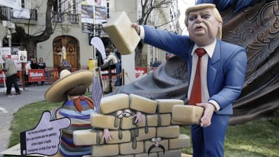 En spansk träskulptur karrikerar Donald Trump som bygger en mur mot Mexiko av tegelstenar. Människor har fastnat mellan murens tegel.