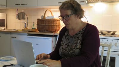 En kvinna sitter i ett kök, vid köksbordet, och jobbar vid en dator.