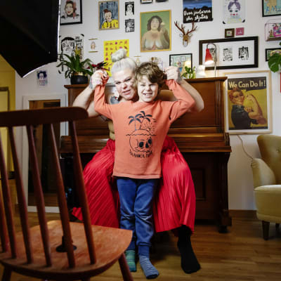 En kvinna sitter på en pianostol och håller en liten pojkes händer upp i luften. Båda grimaserar glatt.