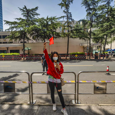 Kvinna med mörkt hår står utanför konsulat och håller upp Kinas flagga. Kvinnan är klädd i röd jacka och bär munskydd.