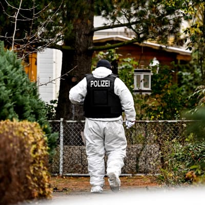 En polis i vit skyddsdräkt och mörk skyddsväst med texten Polizei på ryggen går mellan häckar i ett småhusområde i höstfärger.