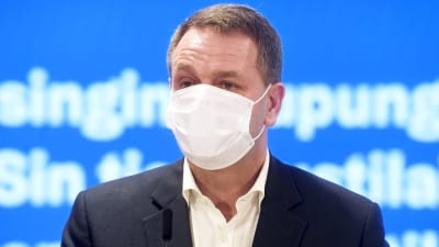 Jan Vapaavuori i munskydd på presskonferensen. Turkos bakgrund.