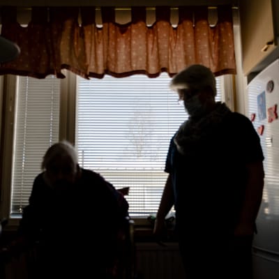 Anonym bild på en äldre och en hemvårdare. Siluetterna syns mot ett fönster.