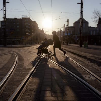 Tampereen Hämeenkatua ylittää rattaita työntävä vanhempi ja pieni lapsi rattaissa. Auringonlasku heittää ihmisistä pitkät varjot tiehen. 