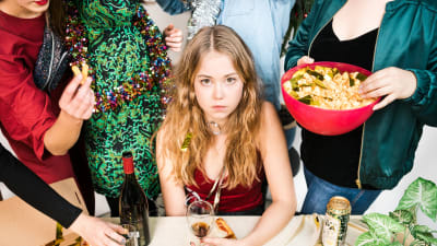 Flicka sitter vid ett bord omgedd av stående kompisar på en hemmafest.