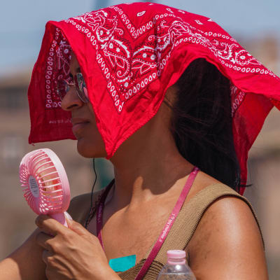En kvinna fotograferad från sidan i närbild. Hon har en röd scarf på huvudet och fläktar sig med en liten fläkt. Hon är klädd i en topp och bär på en flaska vatten.