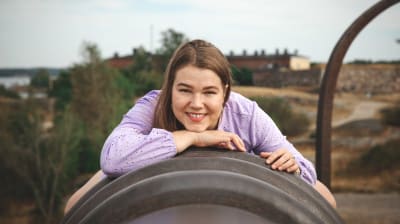 Saana Rasehorn makaa tykin piipun päällä Suomenlinnassa. Hän katsoo kameraan ja hymyilee. Yllään hänellä on violetti mekko. Taustalla näkyy Suomenlinnan muuria ja pieni siivu merta.